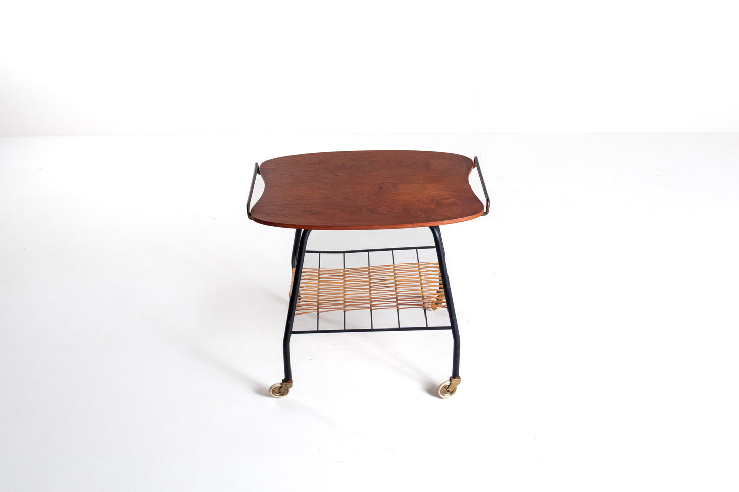 A mid-century modern rolling side table featuring a curved reddish-brown wooden top, a black metal frame, a woven rattan shelf, and brass caster wheels, shown against a plain white background.