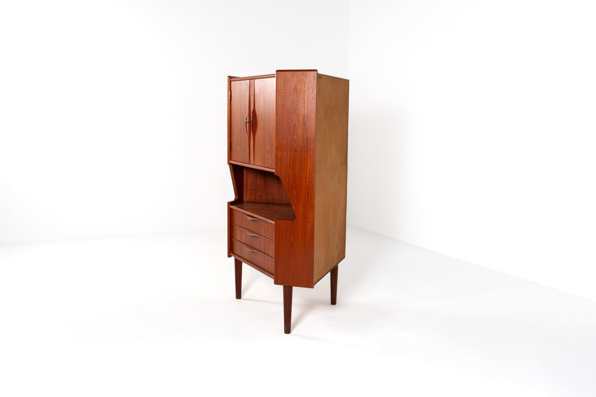 A mid-century modern wooden cabinet with a warm reddish-brown finish, featuring a two-door upper section, an open middle shelf, and three drawers below, set against a plain white studio background.