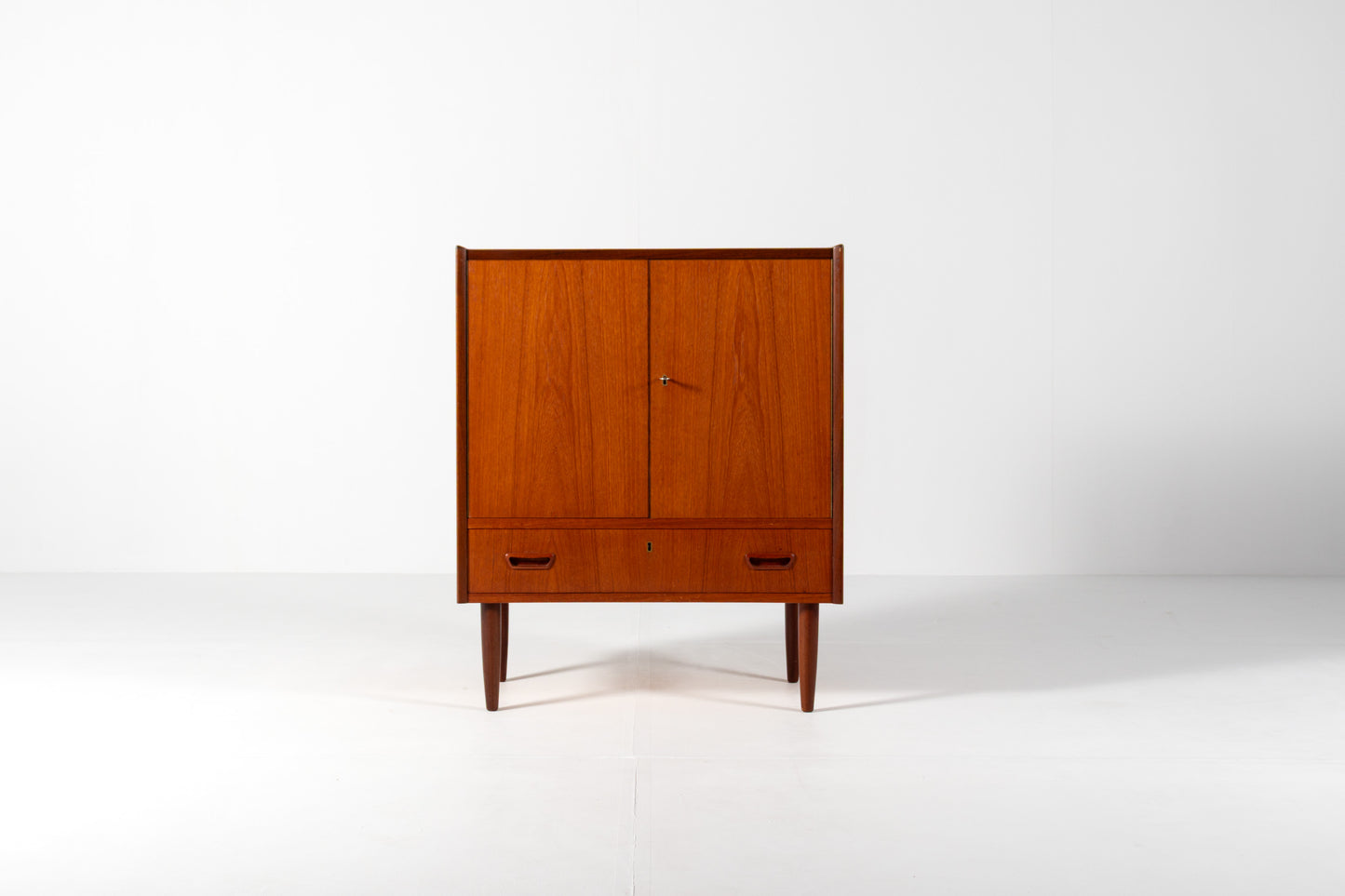 A mid-century modern wooden cabinet with a warm reddish-brown finish, featuring two upper doors and a lower drawer