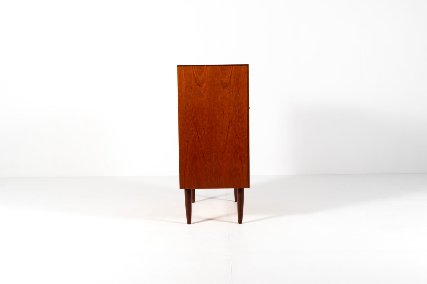 A mid-century modern wooden cabinet with a warm reddish-brown finish, featuring two upper doors and a lower drawer, side view