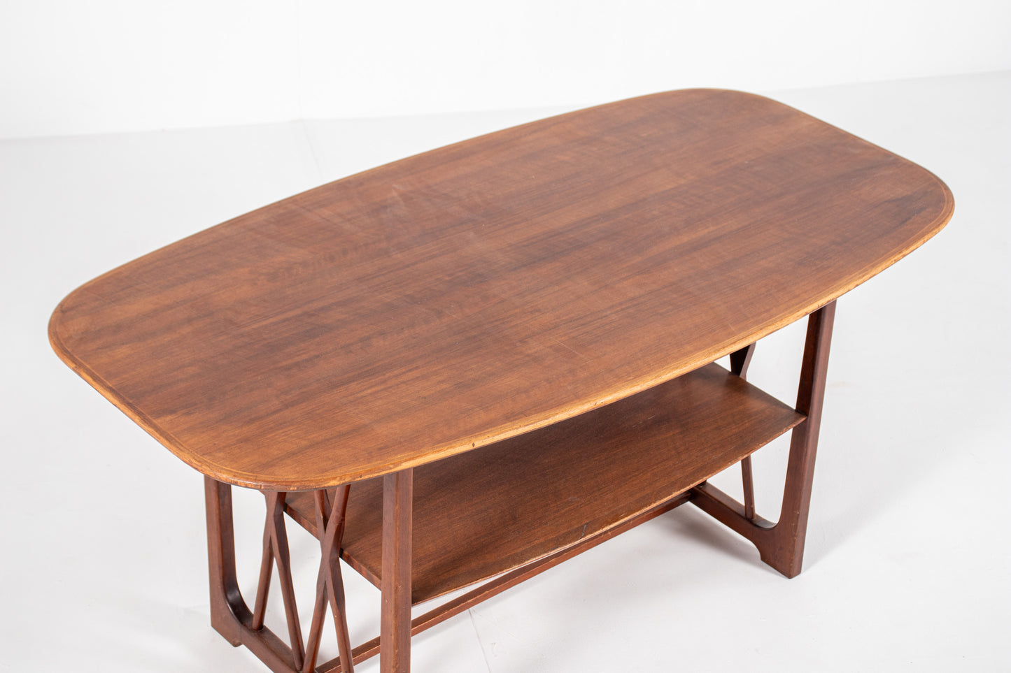 A top of a  mid-century modern wooden table designed by A.A. Patijn for Zijlstra Joure with a warm brown finish, featuring a rounded rectangular top, two open shelves, and geometric cross-brace leg supports, set against a plain white studio background.