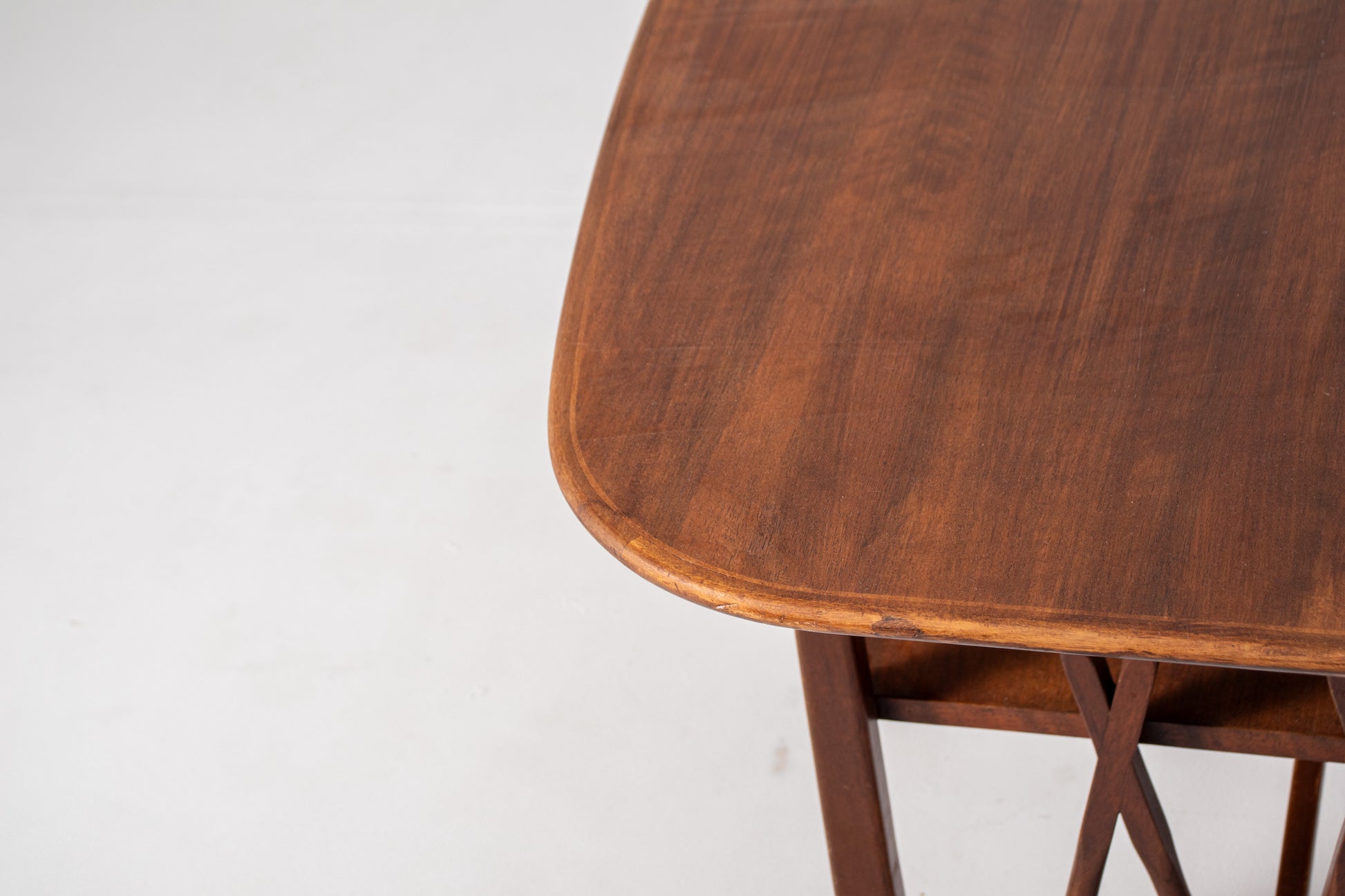A close-up of a  mid-century modern wooden table designed by A.A. Patijn for Zijlstra Joure with a warm brown finish, featuring a rounded rectangular top, two open shelves, and geometric cross-brace leg supports, set against a plain white studio background.