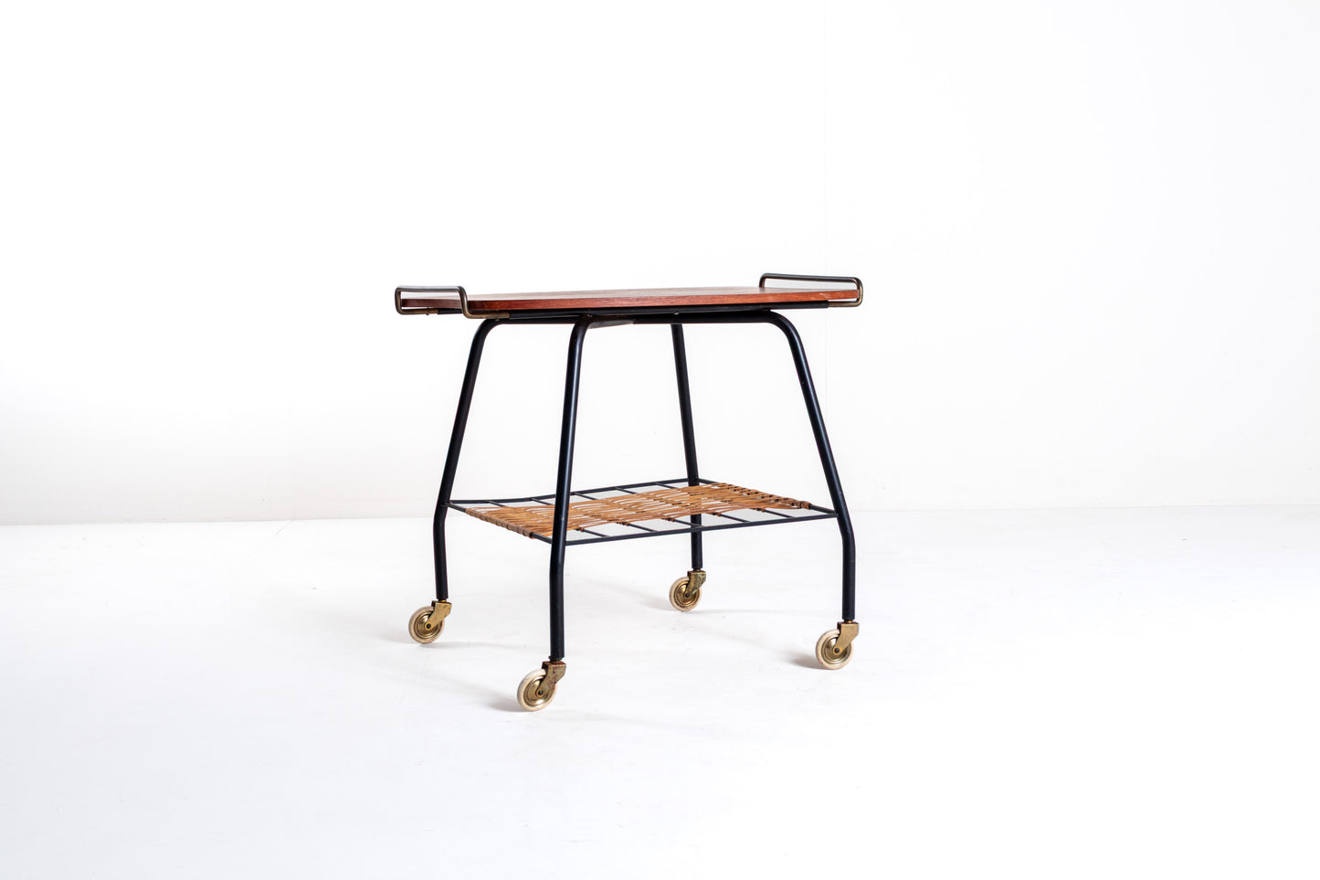 A mid-century modern bar cart with a dark wood top, black metal frame, a slatted wooden lower shelf, and brass caster wheels, photographed on a seamless white studio background.