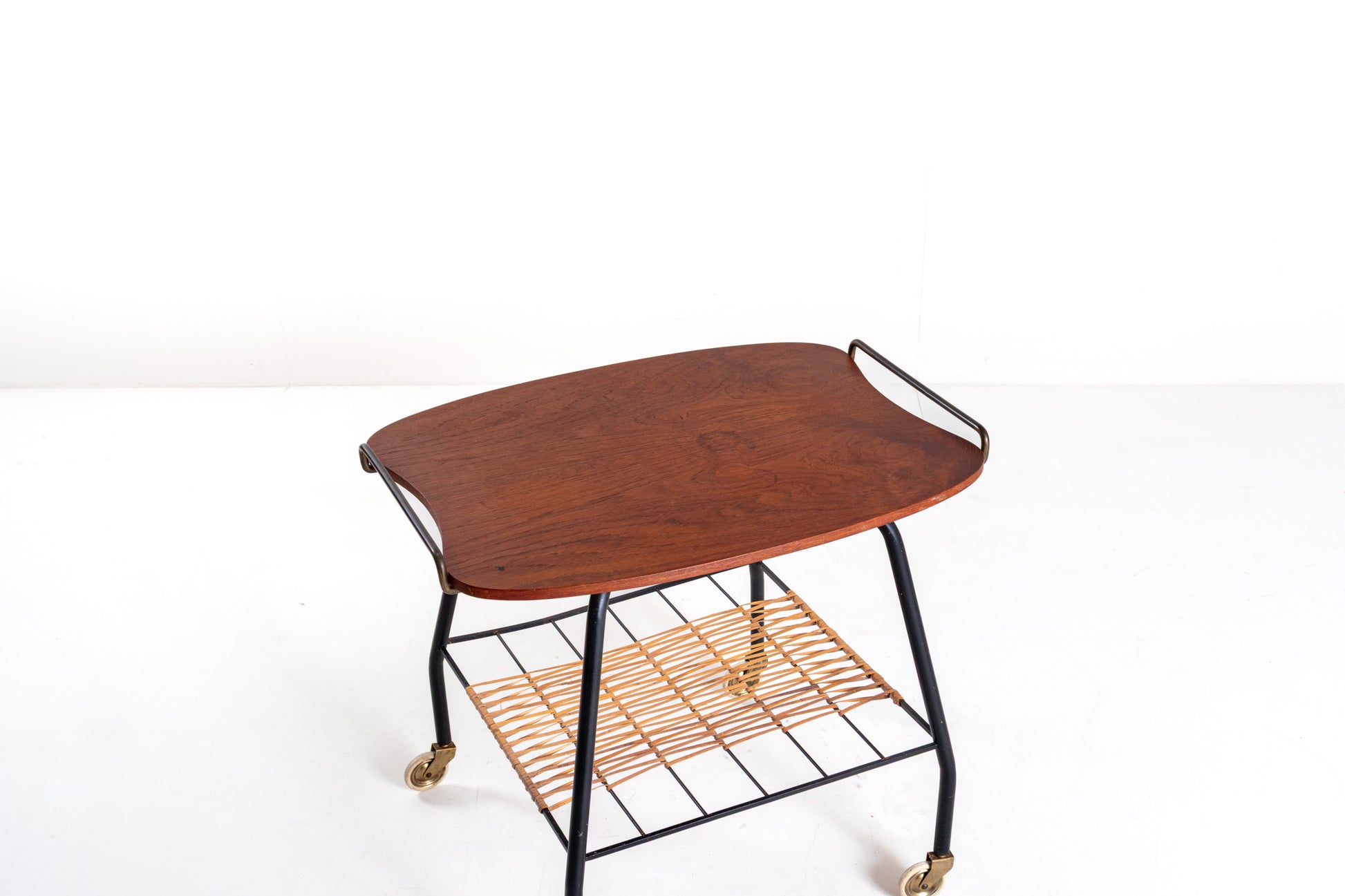 A mid-century modern serving cart with a curved reddish-brown wood top, black metal frame, and a woven rattan lower shelf on casters, photographed against a white studio background.