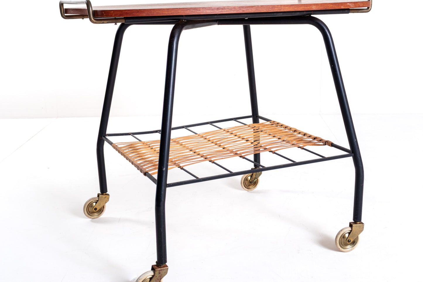 A mid-century modern rolling bar cart with a black metal frame, a warm brown wood-grain top, and a woven tan rattan lower shelf on brass caster wheels.