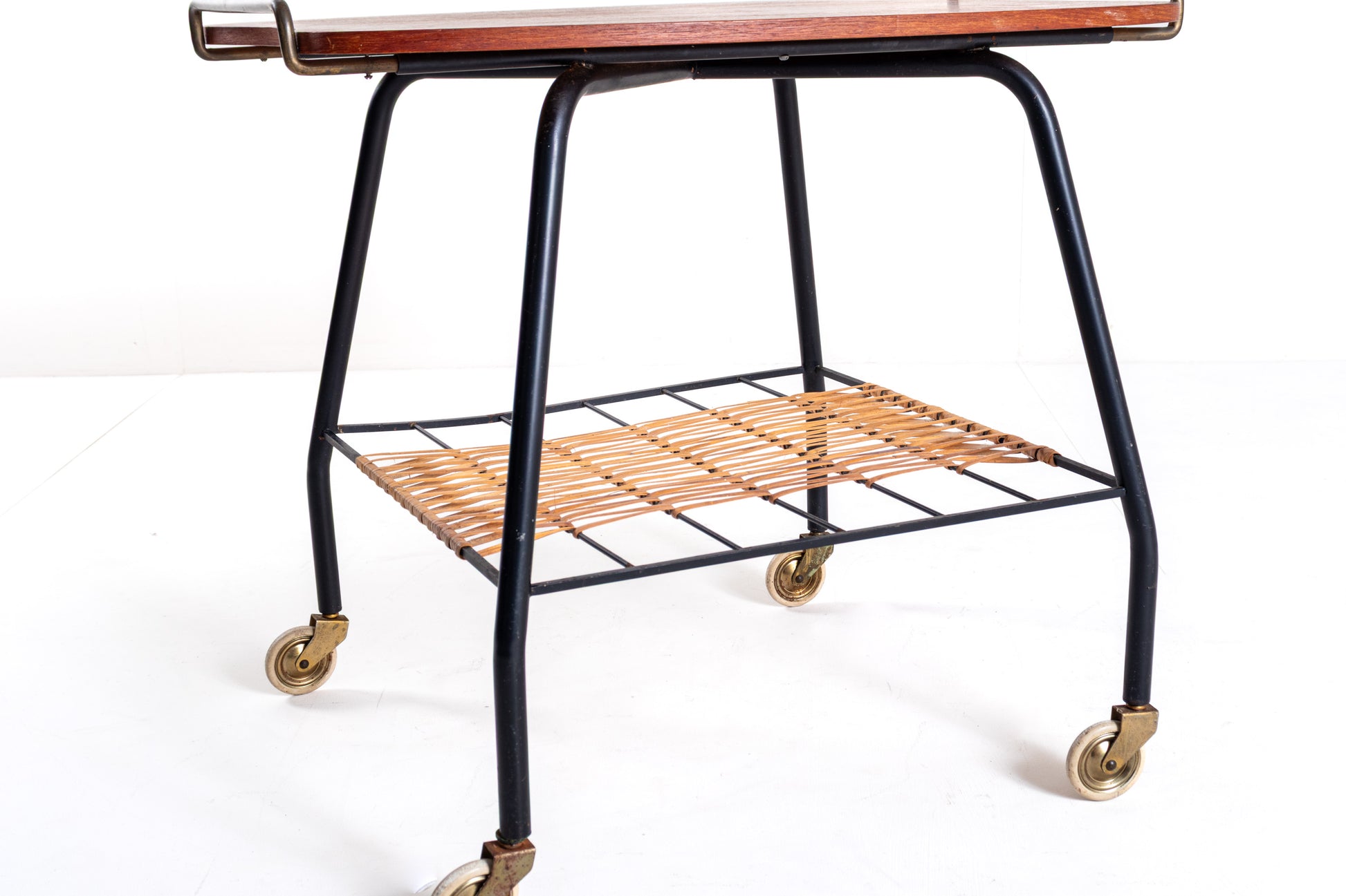 A mid-century modern rolling bar cart with a black metal frame, a warm brown wood-grain top, and a woven tan rattan lower shelf on brass caster wheels.