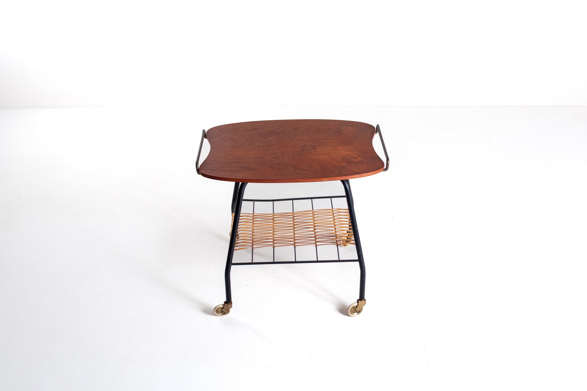 A mid-century modern rolling side table featuring a curved reddish-brown wooden top, a black metal frame, a woven rattan shelf, and brass caster wheels, shown against a plain white background.