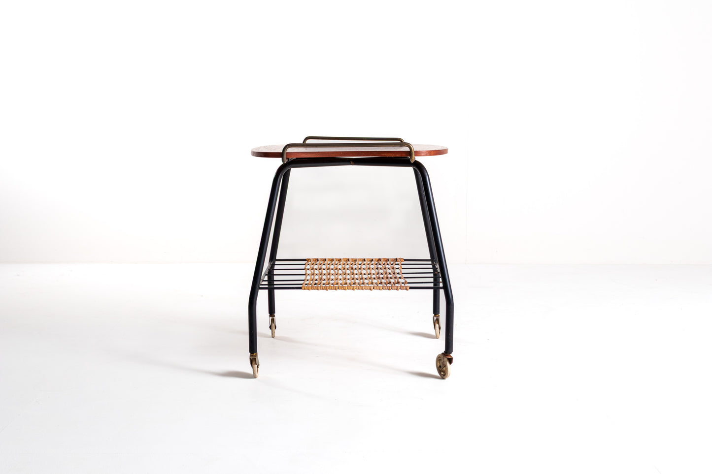 A mid-century modern rolling side table with a black metal frame, a curved reddish-brown wooden top, and a woven rattan shelf, shown on a white studio background.