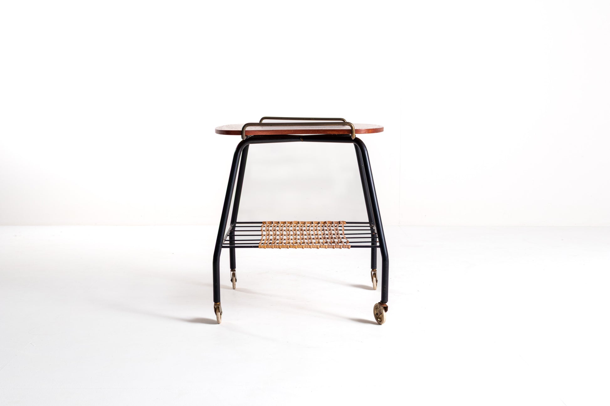 A mid-century modern rolling side table with a black metal frame, a curved reddish-brown wooden top, and a woven rattan shelf, shown on a white studio background.